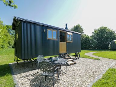 A small black corrugated metal shepherd hut with wooden door and windows on a gravel area with metal table and chairs in a green grassy outdoor setting at Blackthorn South Leigh near Witney