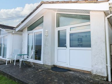 An exterior view of a building with white walls glass doors windows and outdoor chairs with a table on a concrete patio at Chalet 27 in Port Eynon
