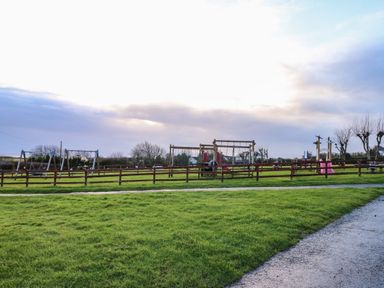 A playground with swings slides and climbing frames surrounded by a wooden fence and grassy field at Chalet 27 in Port Eynon