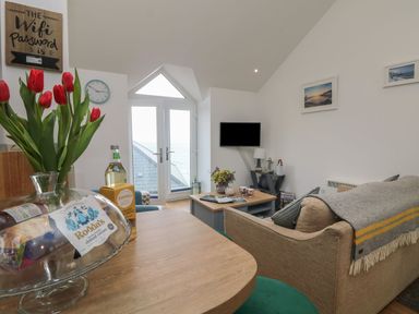 A living room with a beige sofa a wooden coffee table a tv mounted on the wall and glass doors with a sea view at The Loft in Porthleven