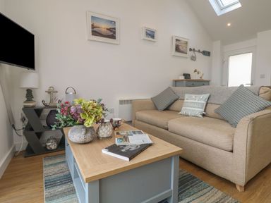 A living room with a beige sofa and cushions a wooden coffee table with flowers a TV on a stand and framed pictures on the wall at The Loft in Porthleven