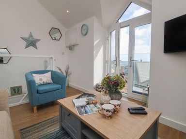 A living room with a blue armchair a wooden coffee table with flowers cups and magazines near a glass door and a wall-mounted TV at The Loft in Porthleven