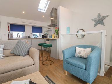 A living area with a beige sofa, blue armchair, bar stools, and a kitchen area at The Loft in Porthleven