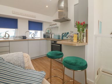 A kitchen with grey cabinets blue window blinds green stools and flowers on a counter at The Loft in Porthleven