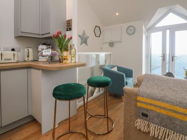 A kitchen area with a wooden countertop a vase of red tulips two green bar stools a blue armchair and a beige sofa with a grey and yellow throw at The Loft in Porthleven