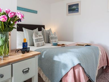 A bedroom with a bed with cushions and blankets next to a bedside table with flowers sunglasses and sunscreen at The Loft in Porthleven
