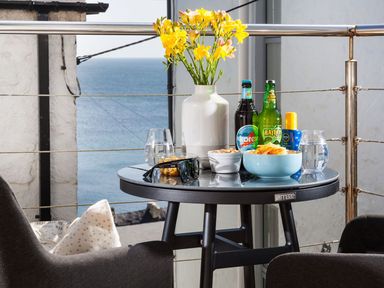 A balcony table with flowers drinks snacks sunglasses and glasses of water overlooking the sea at The Loft in Porthleven