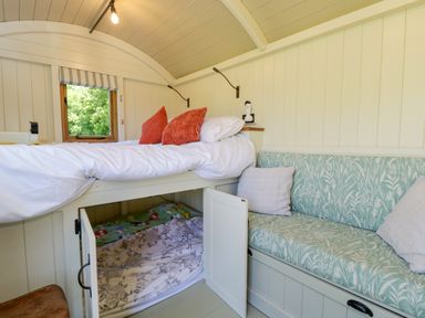 An interior with a raised bed with red pillows a built in bench with patterned cushions and an open cabinet door showing bedding Hazel in South Leigh near Witney