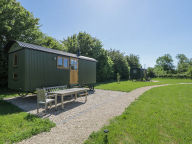 Two green metal shepherd huts with wooden doors on a grassy field with gravel paths and outdoor wooden seating at Hazel in South Leigh near Witney