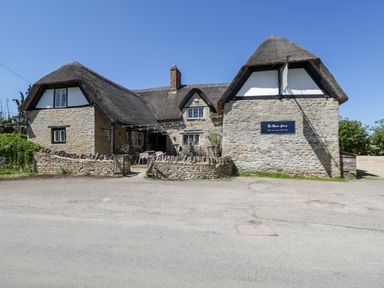 A stone building with a thatched roof and outdoor seating area by a road at Hazel South Leigh near Witney