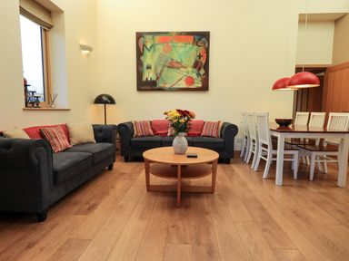 A living room with two dark sofas and a round wooden table with flowers and a dining table with white chairs at Cedrus Lodge in Skelton near Penrith