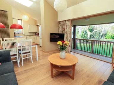 An open living space with a wooden dining table and white chairs a round wooden coffee table with flowers a dark sofa a kitchen area with white cabinets and appliances and a sliding door opening to a green garden at Cedrus Lodge in Skelton near Penrith