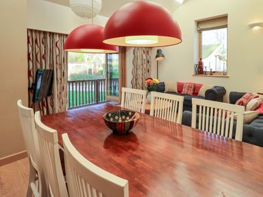 A dining area with a wooden table and white chairs next to a living room with grey sofas and red cushions at Cedrus Lodge in Skelton near Penrith