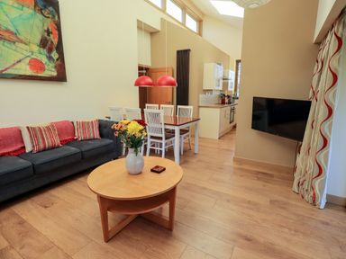 A living room with a dark sofa, wooden coffee table, dining table with white chairs, kitchen area, and a wall-mounted TV at Cedrus Lodge in Skelton near Penrith