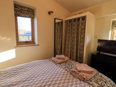 A bedroom with a bed with folded towels a window a tall mirror a TV on a dresser and a curtain covering a closet at Cedrus Lodge in Skelton near Penrith