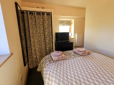 A bedroom with a bed, folded towels, a dresser with a TV, a window with a patterned blind, and a curtain covering a closet at Cedrus Lodge in Skelton near Penrith