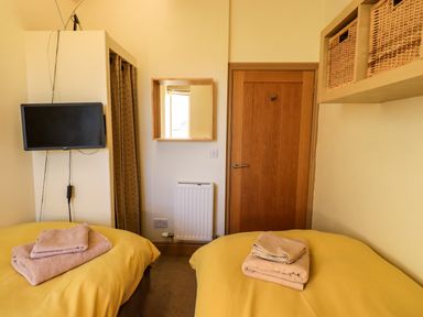 A bedroom with two beds with yellow covers and folded towels on them a wall mounted TV a wooden door and wicker baskets on a shelf at Cedrus Lodge in Skelton near Penrith