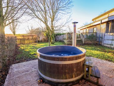 An outdoor hot tub on a concrete patio next to a wooden fence and trees at Cedrus Lodge in Skelton near Penrith