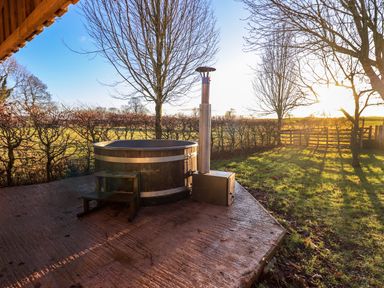 A wooden hot tub with metal bands and a metal stove on a concrete patio in a garden with leafless trees at Cedrus Lodge in Skelton near Penrith