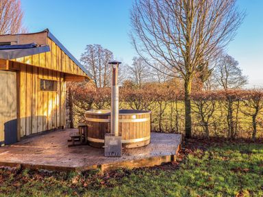 An outdoor wooden hot tub next to a wooden shed on a concrete platform with trees and grass at Cedrus Lodge in Skelton near Penrith