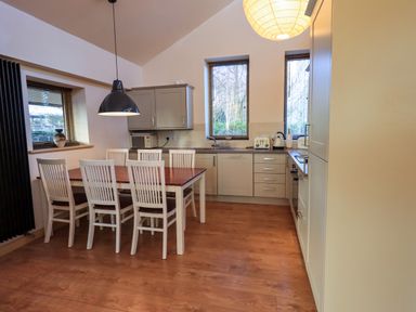 A kitchen with a wooden dining table and six chairs two windows kitchen cupboards and appliances at Larch Lodge in Skelton near Penrith