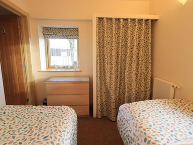 A bedroom with two beds a wooden chest of drawers a window with a blind and a curtain-covered closet at Larch Lodge in Skelton near Penrith