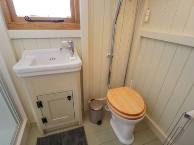 A small bathroom with a white sink cabinet a wooden toilet seat a silver trash can and a gray bath mat at Spindle South Leigh near Witney