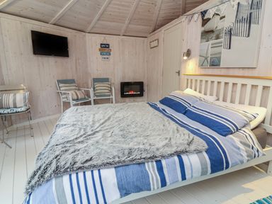 A bedroom with a bed with striped bedding two chairs a wall-mounted television and an electric fireplace at Barley Bay near Hartland