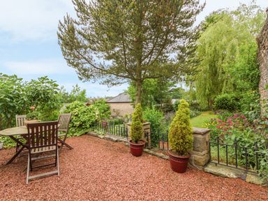 A garden patio with a wooden table and chairs surrounded by plants and trees at Crescent Cottage in Haltwhistle near Hadrians Wall