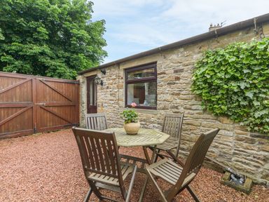 An outdoor patio with a wooden table and four chairs a potted plant stone wall wooden gate and greenery at Crescent Cottage Haltwhistle near Hadrians Wall