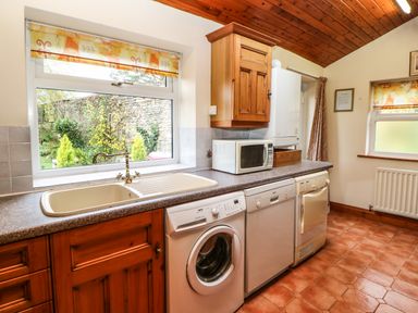 A kitchen with wooden cabinets a washing machine dishwasher microwave and windows showing a garden at Crescent Cottage in Haltwhistle near Hadrians Wall
