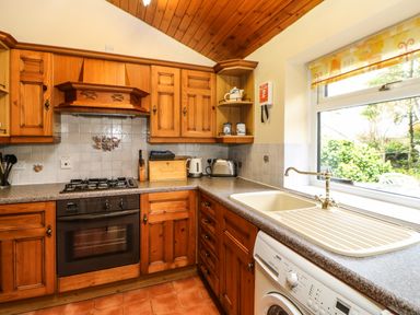 A kitchen with wooden cabinets a gas stove oven a sink a washing machine and a window overlooking greenery at Crescent Cottage in Haltwhistle near Hadrians Wall