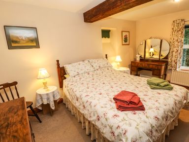 A bedroom with a floral bedspread two bedside tables with lamps a wooden dresser with a mirror and a window with floral curtains at Crescent Cottage in Haltwhistle near Hadrians Wall