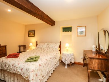 A bedroom with a bed covered in floral bedding two bedside tables with lamps a wooden chair a dresser with a mirror and framed pictures on the walls at Crescent Cottage in Haltwhistle near Hadrians Wall