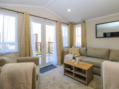 A living room with an armchair and sofa around a wooden coffee table next to French doors and windows with checked curtains at The Sea Lodge in Bacton near Walcott