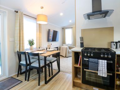 A dining area with a table and four chairs next to a kitchen with a stove and hood and a living room with a sofa and television at The Sea Lodge in Bacton near Walcott