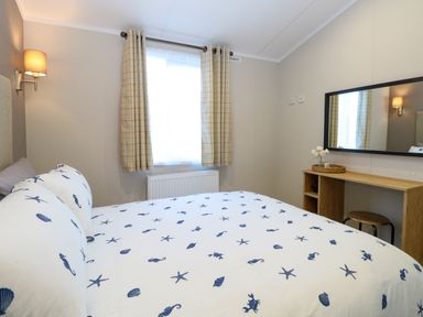A bedroom with a bed with seashell patterned bedding a window with checked curtains a wooden desk with a round stool and a wall mirror at The Sea Lodge in Bacton near Walcott