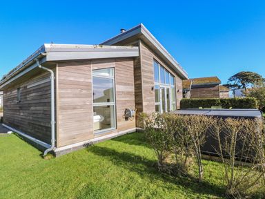 An exterior view of a wooden house with large windows and a covered hot tub on a grassy yard at Una Lodge 77 in Carbis Bay