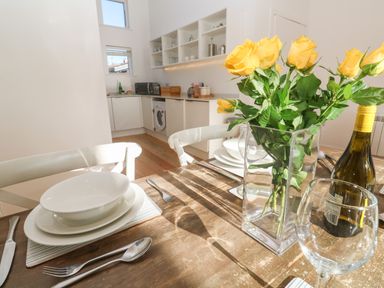 A dining table set with plates bowls cutlery a wine glass a bottle and a vase of yellow roses in a kitchen at Una Lodge 77 in Carbis Bay