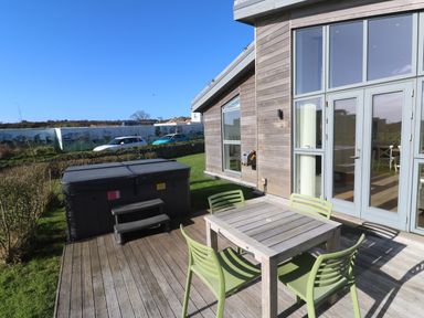 An outdoor wooden deck with a square wooden table and four green chairs next to a hot tub and a modern house with large windows at Una Lodge 77 in Carbis Bay