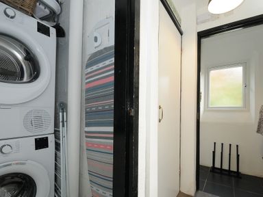 A laundry room with a stacked washer and dryer an ironing board and a small window at Little Ruffo Trebarwith near Tintagel