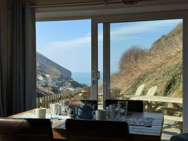 A dining table with cups and glasses by a large window showing a coastal view and outdoor chairs at Little Ruffo in Trebarwith near Tintagel