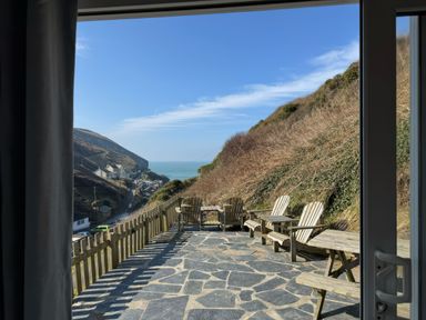 A stone patio with wooden chairs and tables overlooking a hillside and a coastal village at Little Ruffo Trebarwith near Tintagel