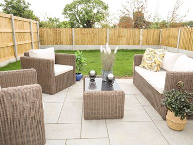 An outdoor patio with wicker seating including chairs and a sofa around a glass coffee table with plants surrounded by a wooden fence at 19 Parc Delfryn in Benllech
