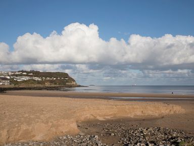 A sandy beach with rocks in the foreground a cliff with buildings on the left and a calm sea under a partly cloudy sky at 19 Parc Delfryn in Benllech