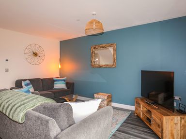 A living room with two gray sofas a glass coffee table a wooden TV stand with a TV a wooden framed mirror blue accent wall and a wall clock at 19 Parc Delfryn in Benllech