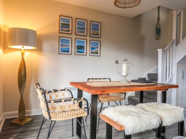 A dining area with a wooden table surrounded by wicker chairs and benches covered with white fur throws a floor lamp and framed pictures on the wall near stairs at 19 Parc Delfryn in Benllech