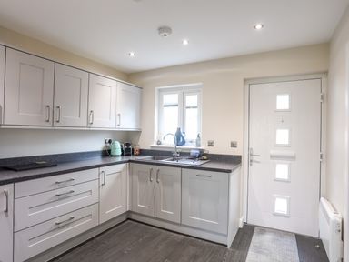 A kitchen with light grey cabinets a white door and a window above the sink at 19 Parc Delfryn in Benllech