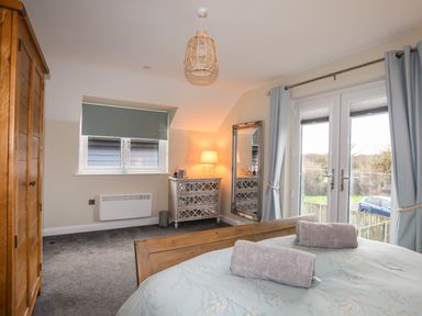 A bedroom with a wooden bed frame grey carpet chest of drawers and large window with curtains at 19 Parc Delfryn in Benllech