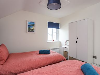 A bedroom with two single beds with red bedding a white wardrobe a small white storage unit a window with a blue blind and a framed picture on the wall at 19 Parc Delfryn in Benllech